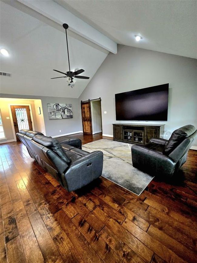 Spacious living room with high vaulted ceilings and recessed lighting. Features rich hardwood floors, a large ceiling fan, and a cozy seating area arranged around a large TV. Includes a sliding barn door and a welcoming entryway. | Image 17