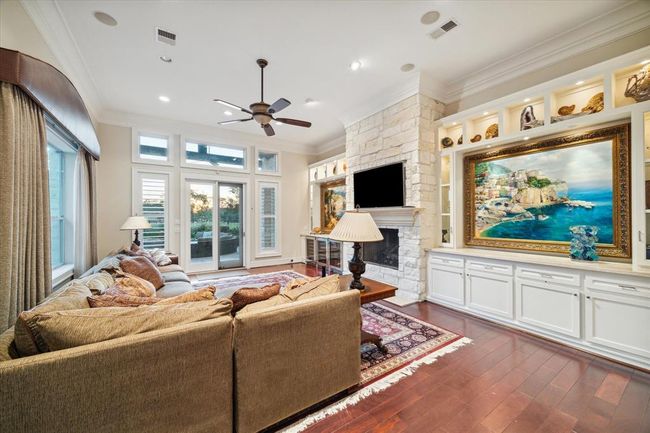 This spacious living room features a stone fireplace, and built-in shelving with tasteful decor. Large windows and French doors provide abundant natural light, leading to a scenic outdoor view. The room is accented with hardwood floors and a ceiling fan, creating a warm and inviting atmosphere. | Image 14