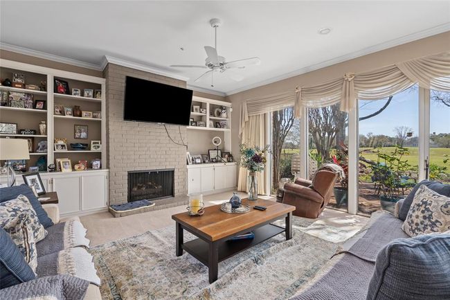 This inviting living space boasts a beautiful brick fireplace, flanked by built-in shelving for storage and display. Floor-to-ceiling windows provide breathtaking views of the lush golf course, filling the room with natural light. The neutral tones and crown molding add a touch of elegance, while the open layout makes it a perfect gathering spot. | Image 11