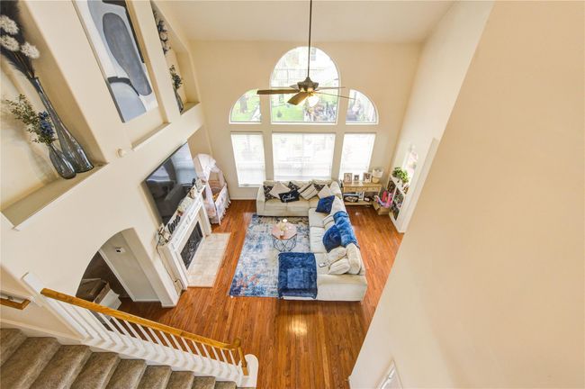 This spacious living room features high ceilings, large arched windows for natural light, and warm wood flooring. It includes a cozy fireplace, a TV, and a comfortable sectional sofa, creating an inviting and elegant atmosphere. | Image 9