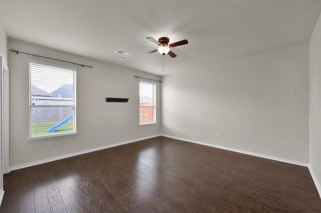 Dark wood flooring and neutral walls. It features a ceiling fan, two doors, and a wall mount for a TV. Large windows provide ample natural light, and there's an entrance to an adjacent bathroom. | Image 17