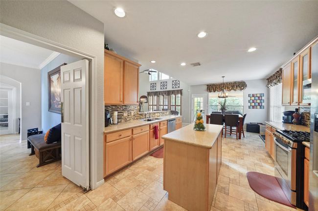 This spacious kitchen features warm wood cabinetry, a central island, and modern stainless steel appliances. The open layout connects to a bright dining area, enhanced by large windows and recessed lighting, creating an inviting atmosphere. | Image 11