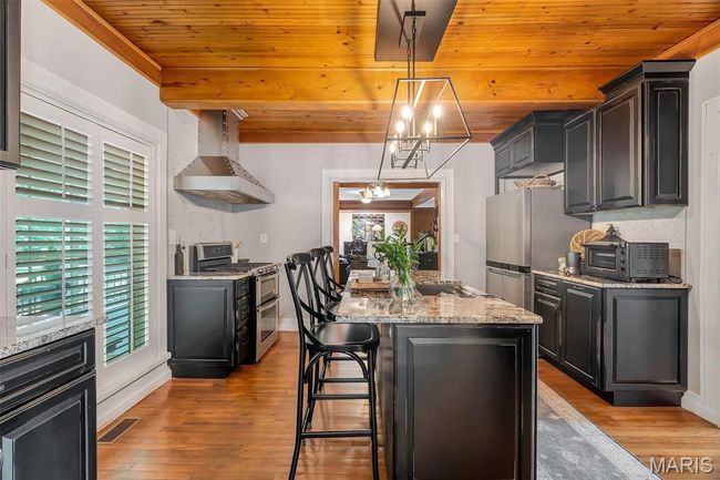 Kitchen featuring wall chimney range hood, appliances with stainless steel finishes, light wood finished floors, wood ceiling, and a center island | Image 46