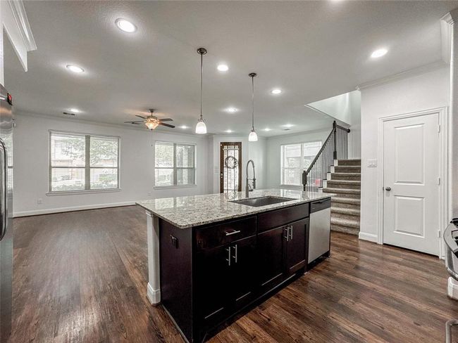This open-concept living space features a modern kitchen with a granite island and dark cabinetry, transitioning into a bright living area with large windows and hardwood floors. A staircase leads to the upper level, and recessed lighting enhances the spacious ambiance. | Image 12