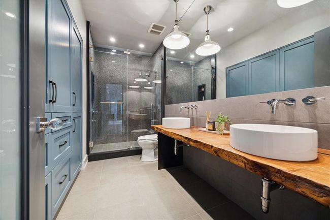 Striking secondary bathroom features a live-edge wood vanity with dual vessel sinks and sleek wall-mounted fixtures. Rich texture is brought in through floor-to-ceiling Thorntree mosaic tile in the walk-in glass shower, adding drama and depth. Custom built-in cabinetry in a deep blue tone provides abundant storage, while industrial pendant lights complete the modern organic aesthetic. A truly memorable and functional space. | Image 33