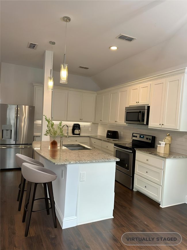 Staged Kitchen featuring stainless steel appliances, vaulted ceiling, backsplash, a center island with sink, and light stone counters | Image 7