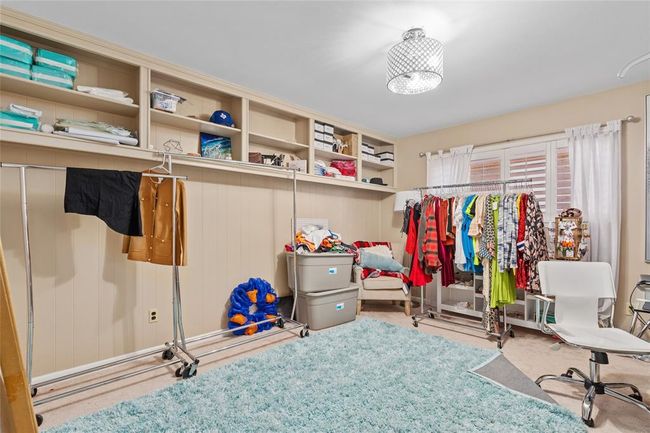 This room features a spacious layout with neutral-toned walls and a light-colored carpet. A large window with plantation shutters allows for ample natural light. Built-in open shelving along one wall provides additional storage, and a ceiling-mounted light fixture brightens the space. | Image 28