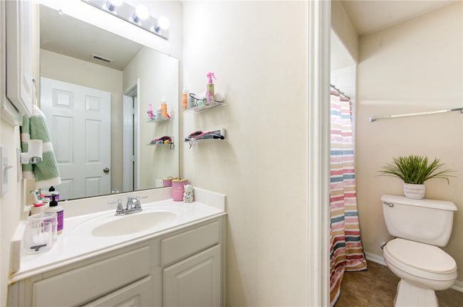 This bathroom features a clean, bright design with a large mirror, ample counter space, and modern lighting. It includes a toilet with a decorative plant, a shower with a colorful curtain, and convenient wall shelving for storage. | Image 32