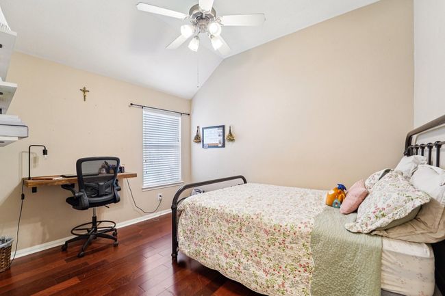 Cozy bedroom with hardwood floors and neutral walls. Features a ceiling fan, a large window for natural light, a simple working desk, and a comfortable bed with floral bedding. Ideal for a peaceful retreat. | Image 18