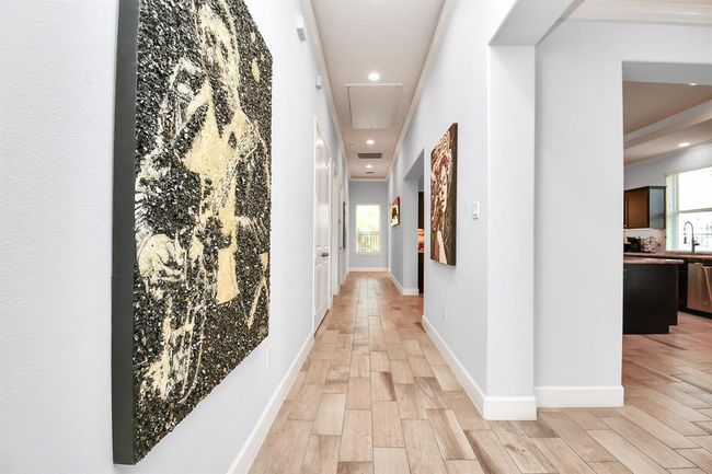 Bright hallway with light wood flooring, modern artwork on the walls, and a view into a spacious kitchen area with ample natural light. | Image 7