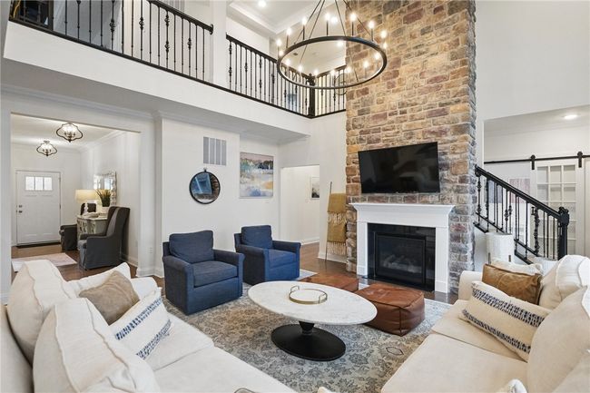 Stunning two-story great room featuring a floor-to-ceiling stone fireplace, dramatic wall of windows with woodland views, and a grand chandelier. Open and airy layout with hardwood floors and abundant natural light, creating a warm and inviting space ideal for relaxing or entertaining. | Image 11