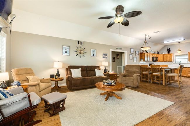 This inviting open-concept living area features a cozy seating arrangement with plush sofas and armchairs, a ceiling fan, and tasteful decor. The space seamlessly flows into a kitchen with warm wooden cabinetry and a breakfast bar, creating a perfect setting for family gatherings. | Image 5
