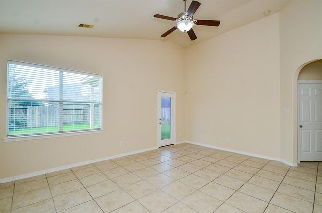 This spacious living room features high ceilings with a ceiling fan, large windows for natural light, tile flooring, and neutral walls. It has a door leading to the backyard, creating a bright and airy living space. | Image 17