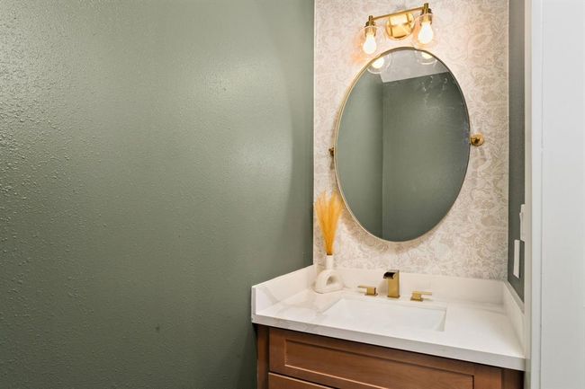 A convenient half bathroom, located near the kitchen, features an elegant vanity with a circular gold pivot mirror and coordinating gold vanity lighting. | Image 35
