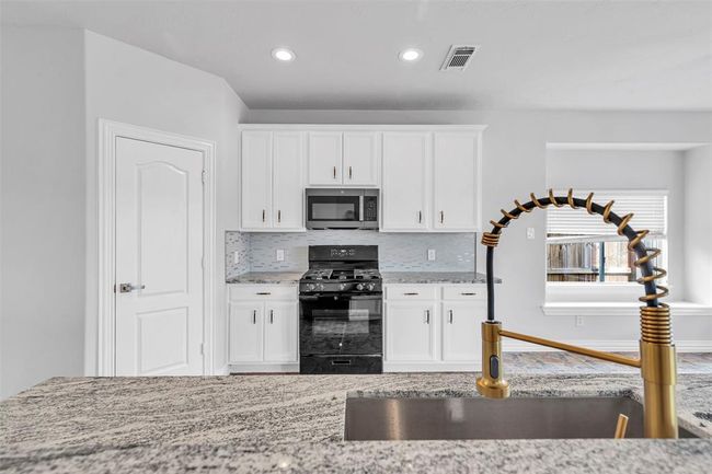 This kitchen features modern white cabinetry, a sleek granite countertop, and a stylish gold faucet. It includes stainless steel appliances, such as a built-in microwave and a gas range, with a light grey backsplash adding a touch of elegance. Natural light streams in through the window, creating a bright and inviting space. | Image 4