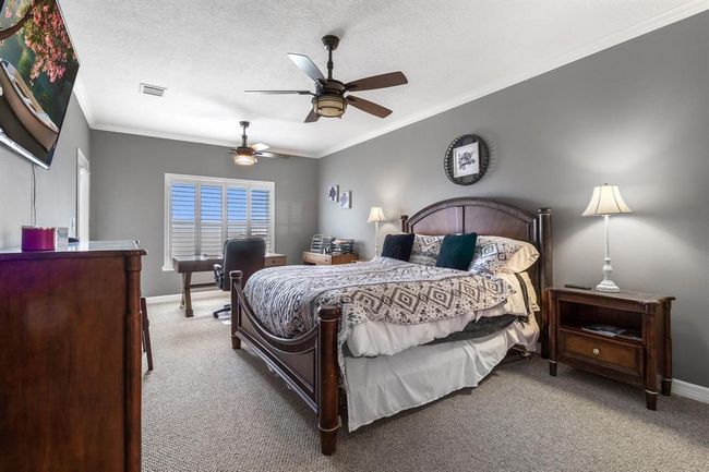 This spacious bedroom features a cozy setup with a large bed, stylish ceiling fans, and elegant grey walls. There's ample natural light from the window with plantation shutters, and the room includes a work desk and classic wood furniture, creating a comfortable and functional space. | Image 14