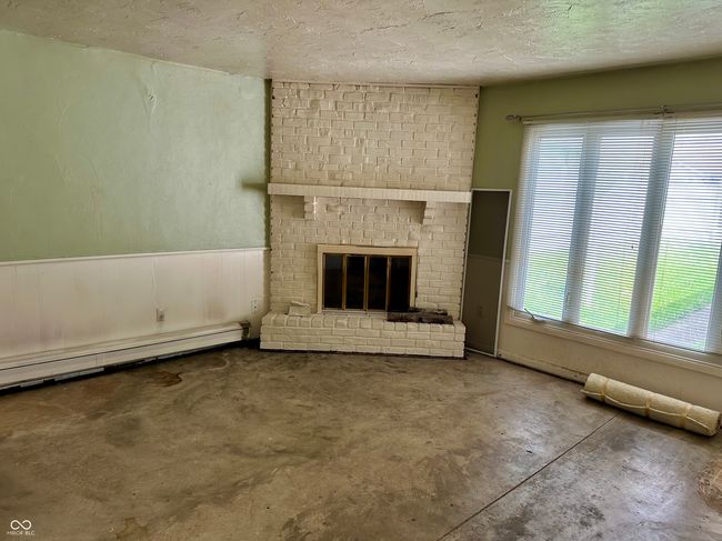 unfurnished living room featuring wainscoting, a baseboard heating unit, a textured ceiling, and a brick fireplace | Image 16