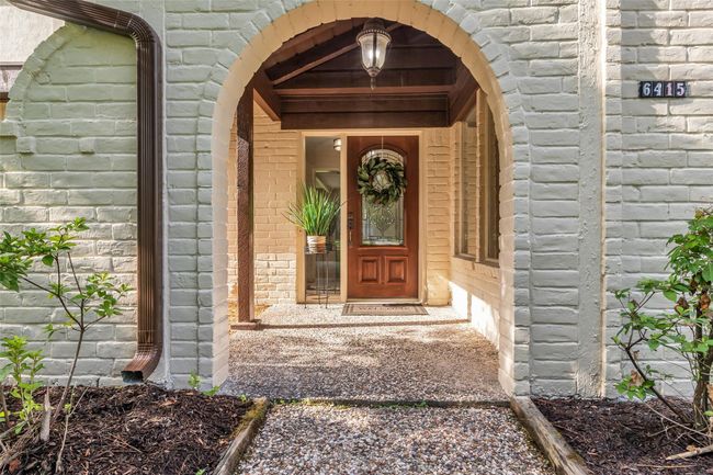 Arched brick accents This inviting entrance features a charming brick archway leading to a wooden front door with decorative glass. The pathway is lined with greenery, and a welcoming light fixture enhances the cozy ambiance. | Image 4