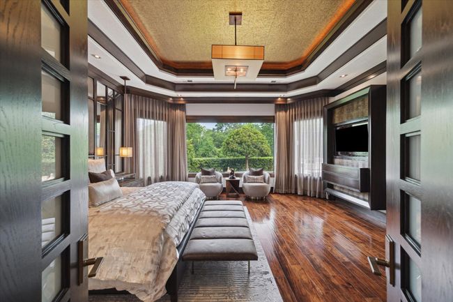 Step through double custom wood and milk glass doors into a refined sanctuary where comfort meets sophistication. Framed by floor-to-ceiling drapery, oversized picture windows bring in tranquil views and soft natural light. The custom tray ceiling with warm metallic inlay and tailored lighting adds architectural interest, while the built-in entertainment center with alligator accents and Musica Net Stream speaker system creates the perfect blend of relaxation and elevated entertainment. | Image 26