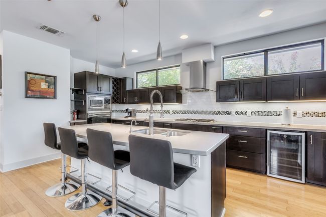 Kitchen featuring wall chimney range hood, stainless steel appliances, wine cooler, hanging light fixtures, and a kitchen island with sink | Image 6