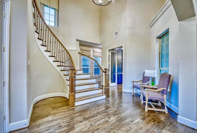 This inviting entryway features elegant hardwood flooring and a striking curved staircase with wrought iron railings. The space is well-lit with natural light and includes a cozy sitting area with two armchairs. The high ceiling adds a sense of openness and sophistication. | Image 9
