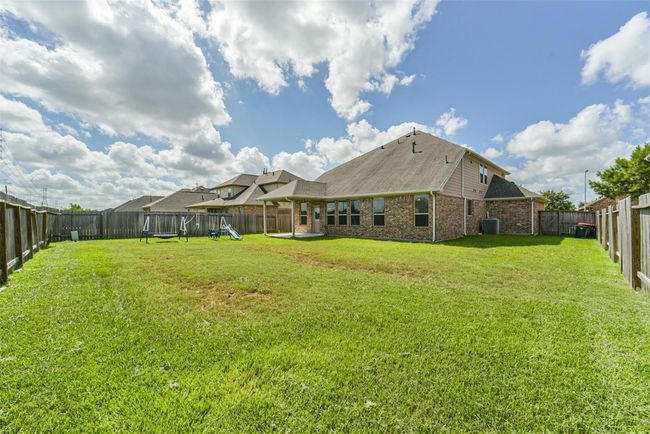 Expansive backyard with lush grass, fenced for privacy, featuring a swing set and a covered patio area attached to a spacious brick house under a clear, partly cloudy sky. Ideal for family activities and outdoor entertaining. | Image 30