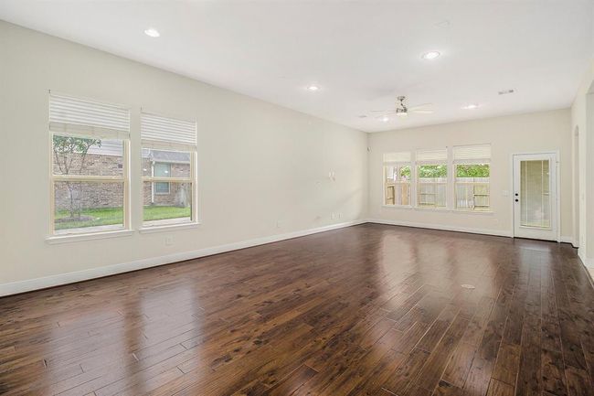 The living room has rich hardwood floors, abundant natural light from large windows, neutral walls, and a ceiling fan. It features a door leading to a fenced backyard, perfect for family living and entertaining. | Image 4