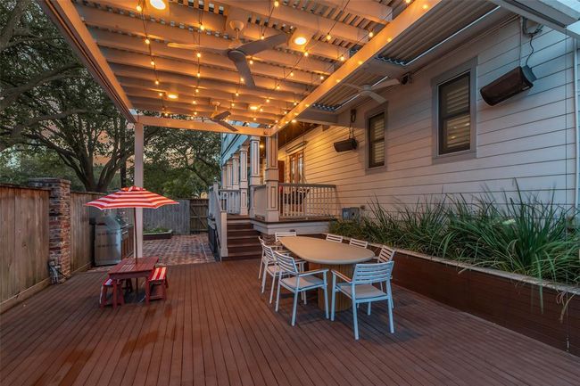 This inviting outdoor dining area with ambient string lighting and ceiling fans. There's a large dining table with chairs, surrounded by lush greenery. An outdoor kitchen completes this oasis! | Image 37