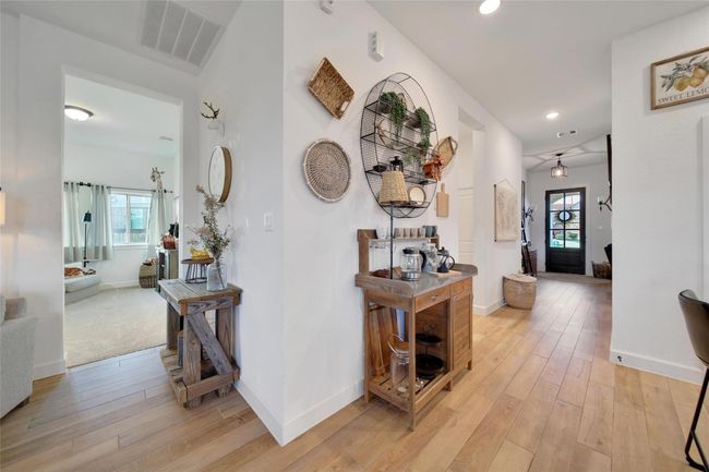 This photo showcases a bright, inviting hallway with wood flooring, leading to a cozy living room on the left. The hallway features a stylish coffee station with rustic décor and a view towards a charming door with glass panels at the end. The space has a modern, farmhouse vibe with thoughtful details. | Image 15