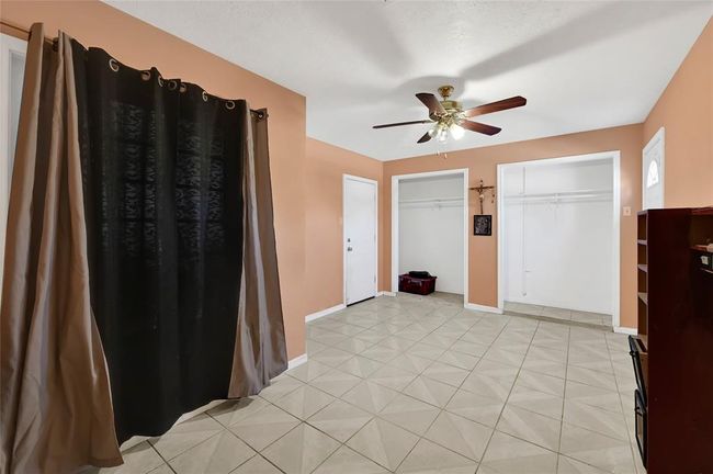 This room features tile flooring, soft peach walls, and a ceiling fan with light. It includes two open closet spaces and a large window with dark curtains, offering a versatile area for multiple uses. | Image 12