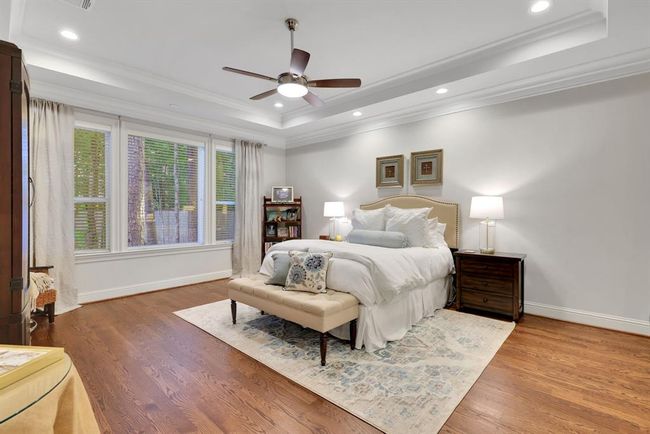 The primary suite is a true retreat—expansive in size and rich in detail. A tray ceiling, paired with double crown moulding, adds depth and character, while warm wood flooring and large windows flood the room with natural light and offer serene views of the park-like backyard. | Image 22
