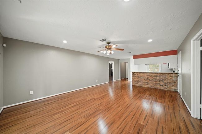 This spacious living area features hardwood vinyl floors and recessed lighting. A brick accent wall adds character, and a ceiling fan provides comfort. The open layout connects to the kitchen, creating a welcoming space. | Image 7