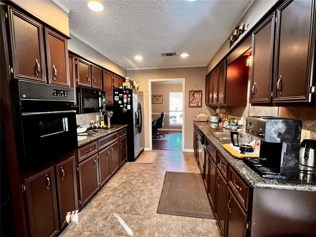 Kitchen with dark brown cabinetry, backsplash, dark stone countertops, a textured ceiling, and black appliances | Image 6