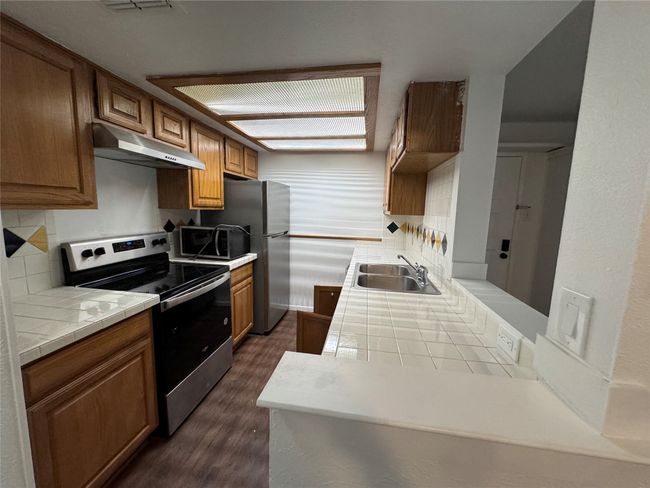 Kitchen with appliances with stainless steel finishes, under cabinet range hood, tile countertops, dark wood finished floors, and brown cabinetry | Image 4