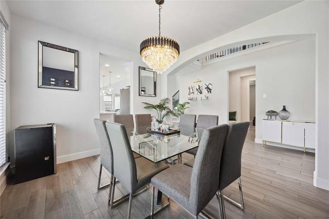 Modern dining area with a glass table and sleek gray chairs, featuring a stylish chandelier. The room is bright and open, with a view into an adjacent space, showcasing contemporary decor and light wood flooring. | Image 6
