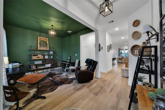 This photo showcases a spacious, open-concept home office with a stylish green accent wall and modern lighting. The space features a cozy seating area, desk, and shelving, with natural wood flooring extending into a bright hallway leading to the living area. Decor elements add warmth and character. | Image 14