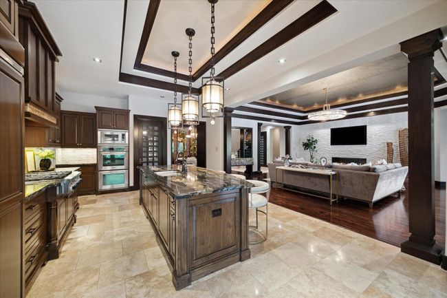 This gourmet kitchen flows effortlessly into the expansive living room, creating a seamless space ideal for everyday comfort. The richly stained cabinetry pairs with exotic granite countertops and a central island that doubles as a roomy gathering spot. Pendant lighting, premium stainless appliances, and tray ceilings with wood accents elevate the overall aesthetic, while the open layout maintains clear sightlines to the fireplace and beyond. | Image 17