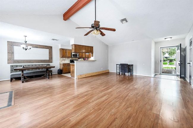 This spacious open-plan living area features high ceilings with a central wooden beam, a ceiling fan, and wood-style flooring. The room flows seamlessly into a cozy dining area. Natural light fills the space through a large window, creating a welcoming atmosphere. | Image 7
