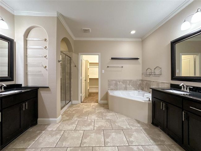 This spacious bathroom features dual vanities with dark wood cabinetry, a large corner bathtub, a glass-enclosed shower, and a walk-in closet, all accented by neutral tiles and soft lighting. | Image 39