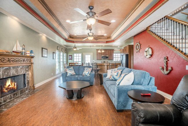Inviting, open living room with hardwood floors and stone fireplace, flowing to kitchen/dining. Features a vibrant red accent wall with nautical décor. Abundant natural light fills the space through well-positioned windows and a rear glass door, enhancing its bright and airy ambiance. | Image 4