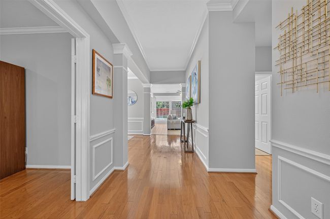 This hallway features elegant wainscoting and crown molding, with warm hardwood floors leading to a bright living area. Natural light enhances the spacious, welcoming atmosphere. | Image 35
