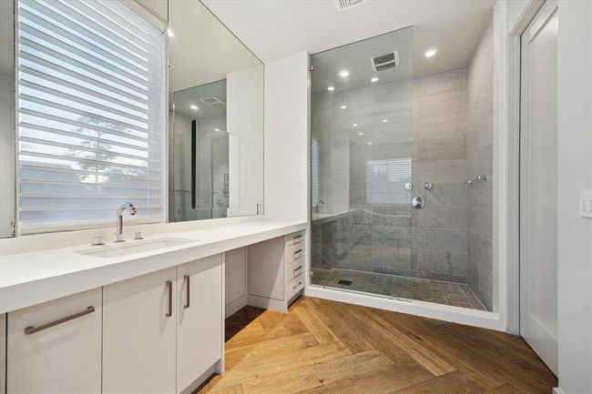 The second side of the primary bath showcases a spacious walk-in shower with floor-to-ceiling tile, seamless glass enclosure, and dual shower heads. A long vanity with under-counter storage and a dedicated seated makeup station offers both style and function. Bright natural light filters through modern window coverings, while warm herringbone wood-look flooring adds elegance and continuity throughout. | Image 18