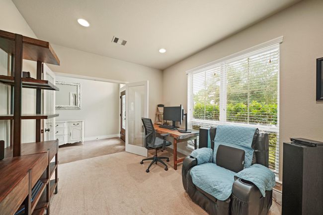 This room is a bright, well-lit office space with a large window, neutral walls, carpeted floor, and French doors, making it ideal for working from home. | Image 8