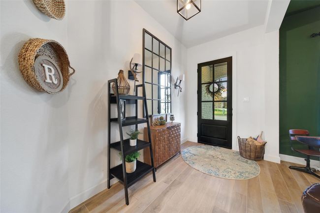 This welcoming entryway features a stylish black door with glass panels, light wood flooring, and a decorative wall mirror. A ladder-style shelf and a woven basket add charm, while a modern light fixture enhances the space. | Image 12