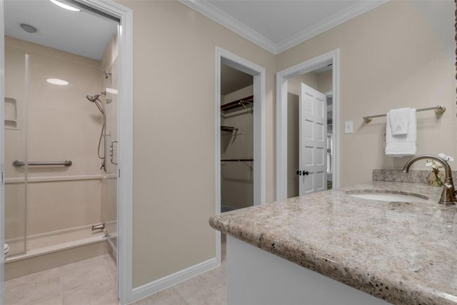 This primary suite bathroom features a walk-in shower with a glass door, a spacious granite countertop with an undermount sink, and neutral tile flooring. Adjacent is a walk-in closet, offering ample storage space. | Image 27