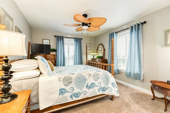 3rd bedroom with beach-themed decor, featuring a ceiling fan, light blue curtains, and a comfortable bed. The room is bright and inviting, with two windows providing ample natural light. | Image 18