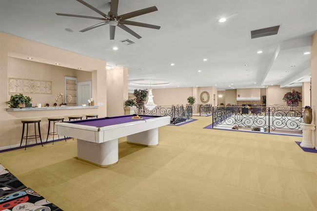 You're looking at a spacious entertainment room featuring a pool table, a bar area with stools, and elegant wrought-iron railings. The room has recessed lighting and a large ceiling fan, providing a comfortable and inviting space for leisure and social gatherings. | Image 21