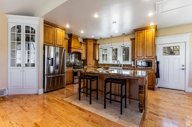 Kitchen with light wood-type flooring, stainless steel appliances, a breakfast bar, brown cabinetry, and custom exhaust hood | Image 9