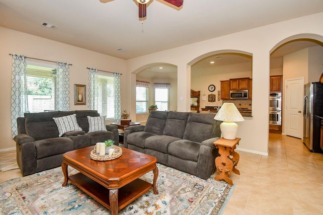 The living room is brightened by natural light from large windows with patterned curtains. It seamlessly flows into the open kitchen, visible through elegant archways, highlighting warm wood cabinetry and modern appliances. | Image 9