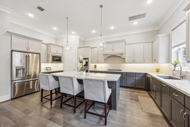 This modern kitchen features sleek stainless steel appliances, a large central island with seating, elegant countertops, and ample cabinetry. Pendant lighting and a neutral color palette create a bright, inviting atmosphere. | Image 12